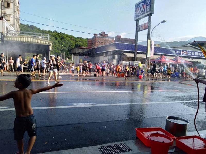 一位赤裸上半身的小男孩打開雙手站在街道邊，身旁是一桶又一桶的水，以及正在向對街灑水的人。