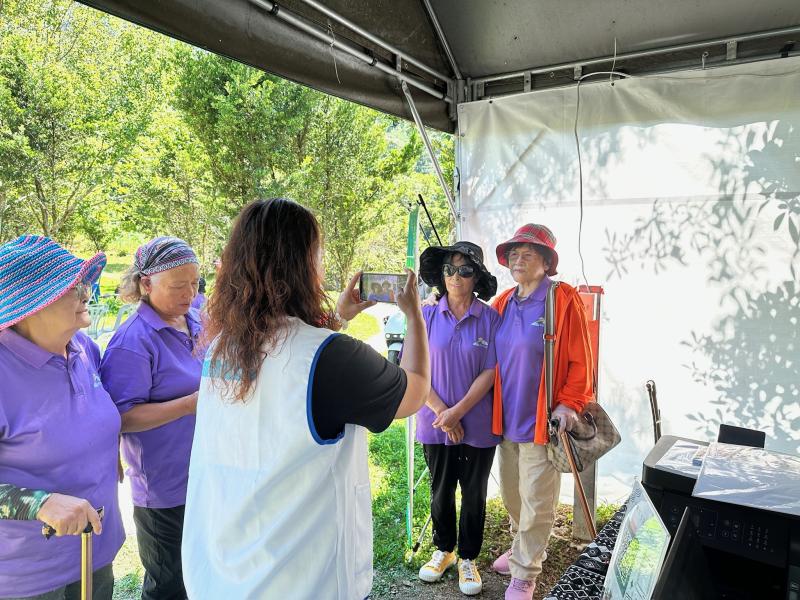 數位中心的攤位引導村民練習用平板電腦拍照。透過 Fotor AI 一鍵生成可愛圖，將在地文化與科技結合。數位幫手協助調整後製，讓原本不熟悉科技的長輩也能輕鬆體驗數位樂趣，操作簡單且充滿互動性。