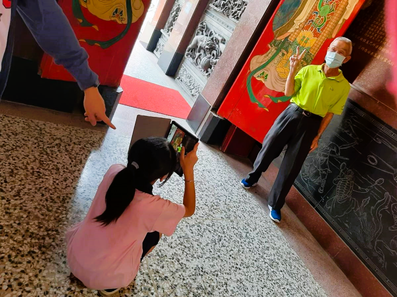 學生在廟前蹲低身姿,手持平板為開心比耶的長輩拍攝人像照,互動溫馨。