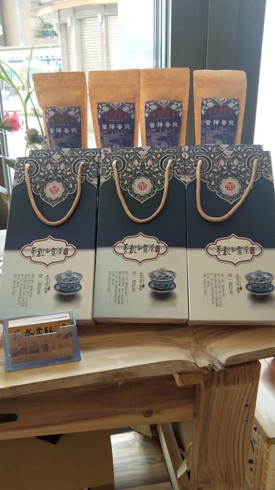 以精美黑色花紋盒裝與橘色袋裝的養氣活靈茶陳列於桌上，外包裝盒上以青花瓷般的茶具作為樣品展示。