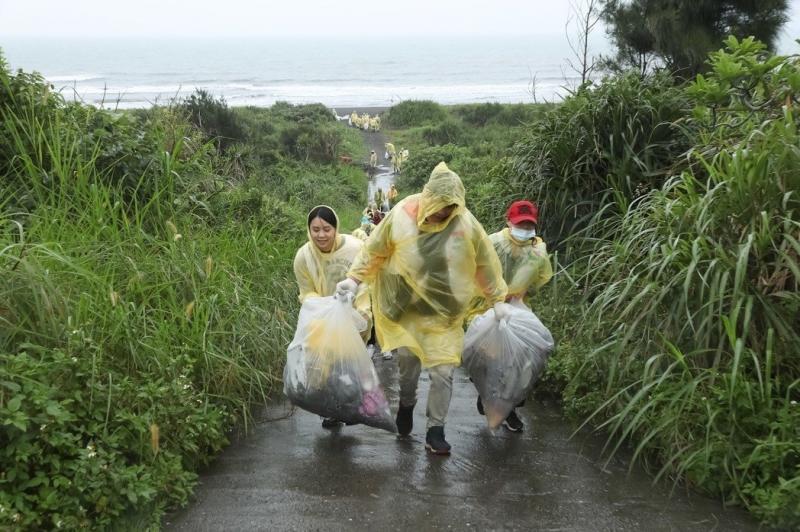 三名旅行志工身穿輕便雨衣，將淨灘結束的垃圾以雙手提起，準備帶離海邊。