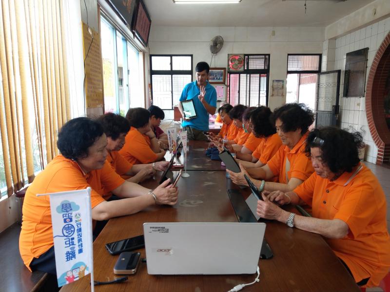 學員們將雙手倚靠於桌，雙眼緊盯近用計劃提供的平板，桌上擺放著旗幟與筆電，講師則站在桌前手抱平板解說。