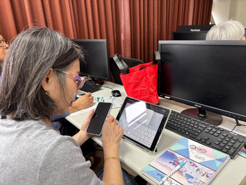 一名女性學員坐在電腦桌前，左手拿著手機，右手正點擊平板電腦螢幕進行註冊與設定練習。桌上放著筆記本，展現出學員在室內課堂上實際操作數位設備的細節。