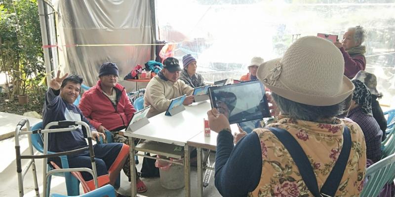 長輩高興的拿著平板，運用剛剛學會的拍照技巧，幫坐在桌前的擺好pose的學員拍照。