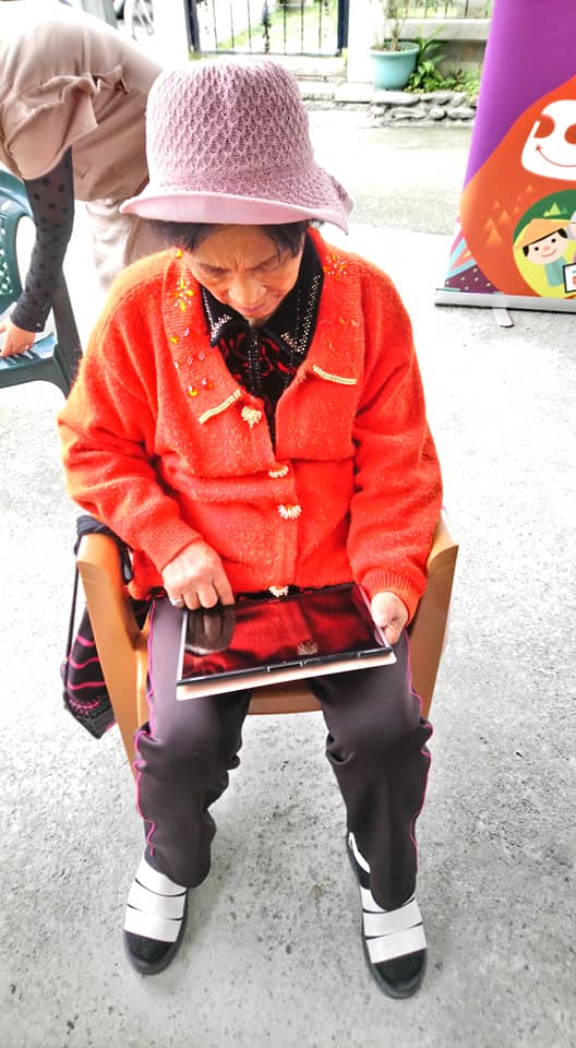 一位穿著紅衣的長輩，將平板放置於雙腿上，用手指緩慢的在平板上滑動。