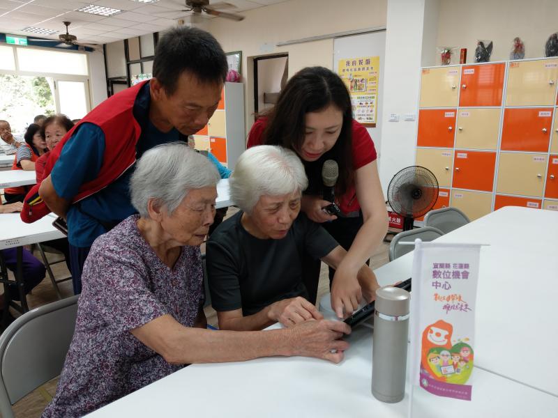 講師立於學員的左方，以手指直接比畫於平板電腦上，兩位長輩學員露出驚訝的表情，似乎學到新事物。