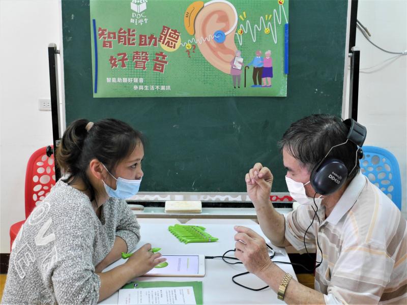 駐點人員(左邊)在桌前給學員(右邊)戴上耳機使用平板進行聽剪