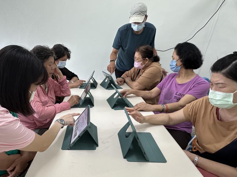 這張根本是訂房大作戰！大家低頭滑平板，有人還喊「欸欸我找到更便宜的欸！」氣氛超級熱絡～老師跟志工忙著一對一指導，長輩們學得超開心。等暑假過後，我們就一起帶著這堂課的成果，去台北住好住滿！