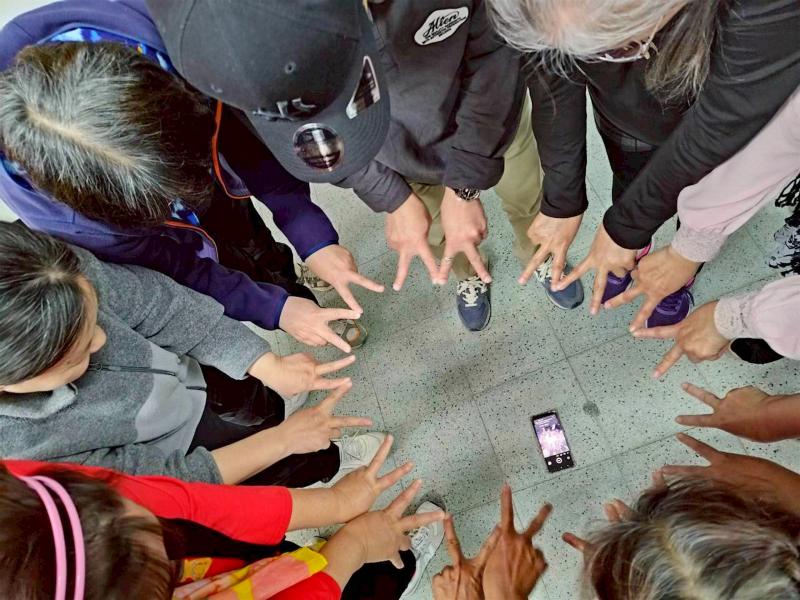 學員們興奮地圍成一個緊密的大圓圈,將手機平放在地板上,開啟前鏡頭與倒數計時器,熱情地伸出雙手比出「YA」的勝利手勢。