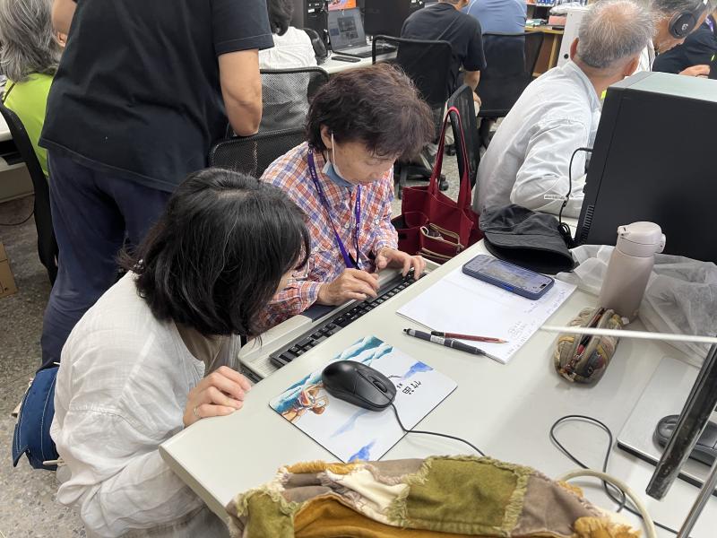 課程中不乏年長的學員，對鍵盤輸入與介面操作較為陌生，輔導員耐心蹲低身子，逐步引導學員完成文字輸入與工具操作。可以看出學員已認真預備好自己想介紹的新埔景點，正一點一點將記憶中的地方故事化為數位作品。