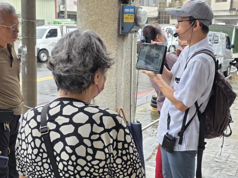 學員們撐著傘、圍繞在講師身旁，一邊聆聽導覽，一邊透過數位螢幕對比今昔地景。