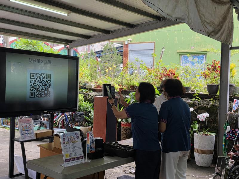 學員於大型電視螢幕前，舉起手機對準螢幕上的防詐大挑戰 QR Code 進行掃描。