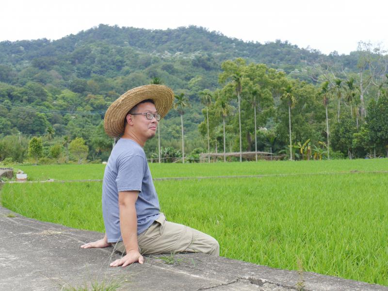 彥同坐在自己的稻田邊，帶著草帽穿著休閒
