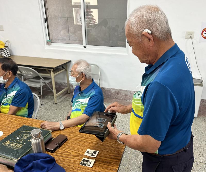 長者專注地使用平板電腦拍攝舊照片，旁邊桌上還擺放著幾張泛黃的舊照片，充滿時光的痕跡。