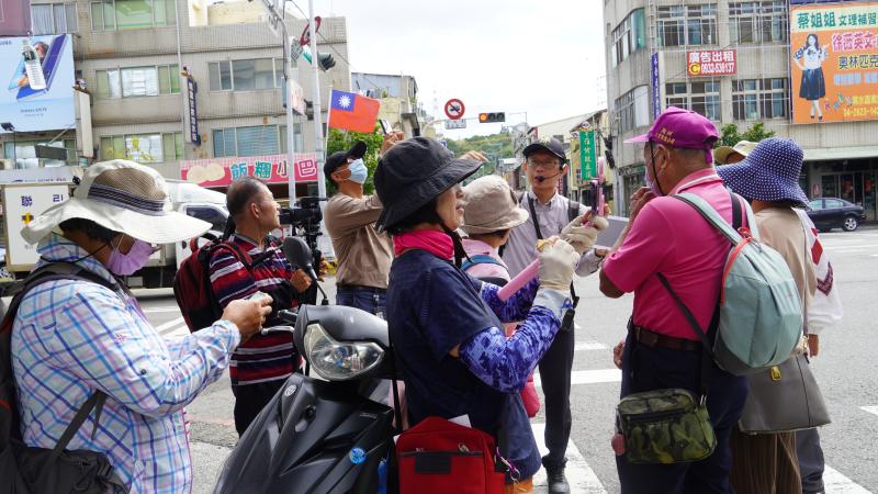 這張照片呈現長輩們在導覽老師的帶領下，以「走讀＋資訊學習」的方式探索清水街區的精彩時刻，透過手中的手機、平板與數位攝影機，他們一邊聆聽在地故事，一邊實際操作拍攝與紀錄，學習如何運用數位工具捕捉地方文化的脈動。老師以即時講述結合地理定位與影像