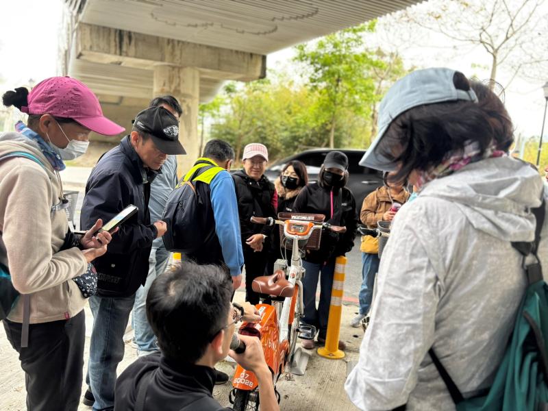 講師蹲在一輛橘色的 YouBike 2.0 電輔車旁,向身邊的學員們現場示範單車檢查要點。多位學員正近距離觀察並認真聽講。