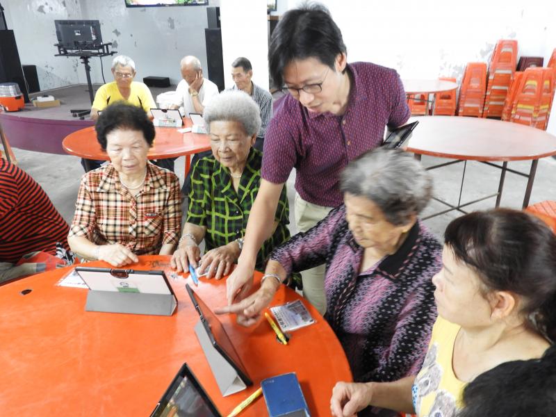 學員們圍坐紅色大桌，並且將平板立起，講師在旁引導長輩由玩小遊戲以熟悉平板。