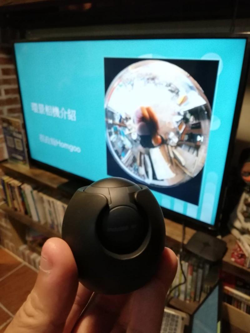 講師將相機以指頭捧著，相機像是一顆黑色的雞蛋外面包覆一層保護殼，中間鏡頭是一個小正圓形。