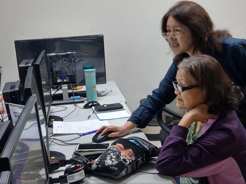 講師站在學員的右方，右手握住學員的滑鼠在螢幕上游移，學員單手托著自己的臉頰雙眼跟隨滑鼠移動。