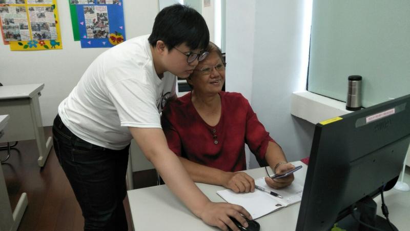 講師手握滑鼠，嘗試解決一位長輩學員的設定問題，學員在一旁拿起手機記錄過程。