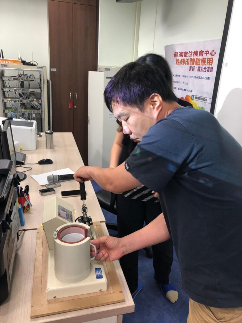 老師左手握住機器的握把，右手將馬克杯放入C型模具中，親自操作熱轉印機器示範給學員看。