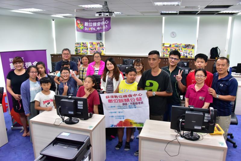 其中一位學員拿起數位智慧公益的課程海報，與講師及學員全體在教室的桌子間合照。