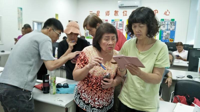 一位學員將手機拿起，詢問其他學員關於使用上的問題，一旁的人認真查看他的問題所在。