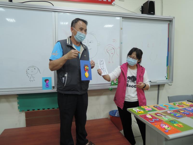 一位男同學在講台上展示自己黏土的作品與手繪的自畫像，老師站在他的旁邊