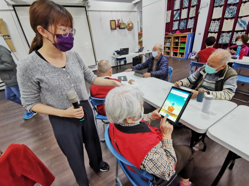 長輩學員獨立操作完成自己的健康飲水計畫任務，使育成的植物成長開花了，正拿著平板在跟老師炫耀