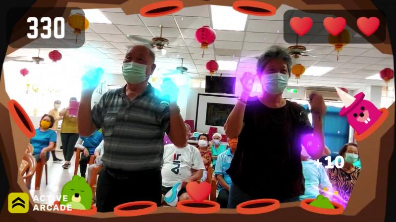 長者們手持設備，跟隨遊戲指引進行各種運動動作，並且積極地參與到遊戲中，臉上洋溢著歡笑和認真的表情。