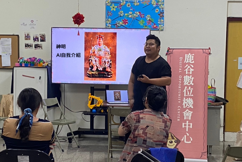 講師正在介紹AI工具如何應用於文化遺產的數位化保存，通過AI技術讓廟宇神像“自己說故事”。在投影屏幕上顯示的是一個鹿谷鄉在地的民間神明慚愧祖師的自我介紹，學員們正認真聆聽，了解如何使用AI技術來活化和保存本地文化。