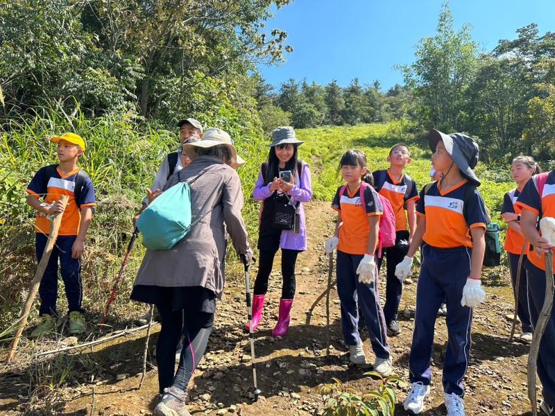 社區長輩與國小六年級學生展開了一場充滿互動的山林探索之旅。照片中可見專業導覽員正熱情解說，讓孩子們透過實地觀察學習古道的自然與文化價值。