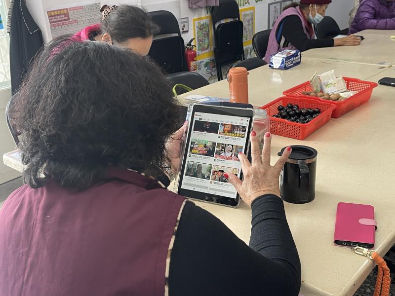 學員正透過平板操作YouTube，搜尋卡拉OK伴奏歌曲，實際練習課堂所學的數位點歌技巧。長者專注於螢幕，展現對新技術的學習興趣，讓歡唱變得更便利。桌上擺放點心，營造輕鬆愉快的學習氛圍。