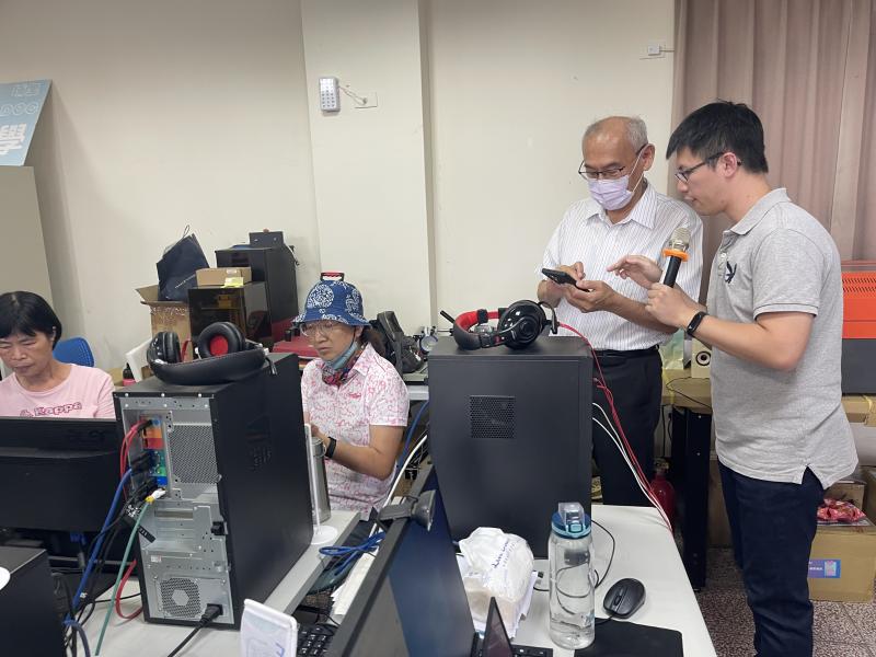 一位長輩專注盯著手機螢幕,身旁的老師一手拿著麥克風、一手比劃操作步驟,耐心地協助他逐步完成任務。