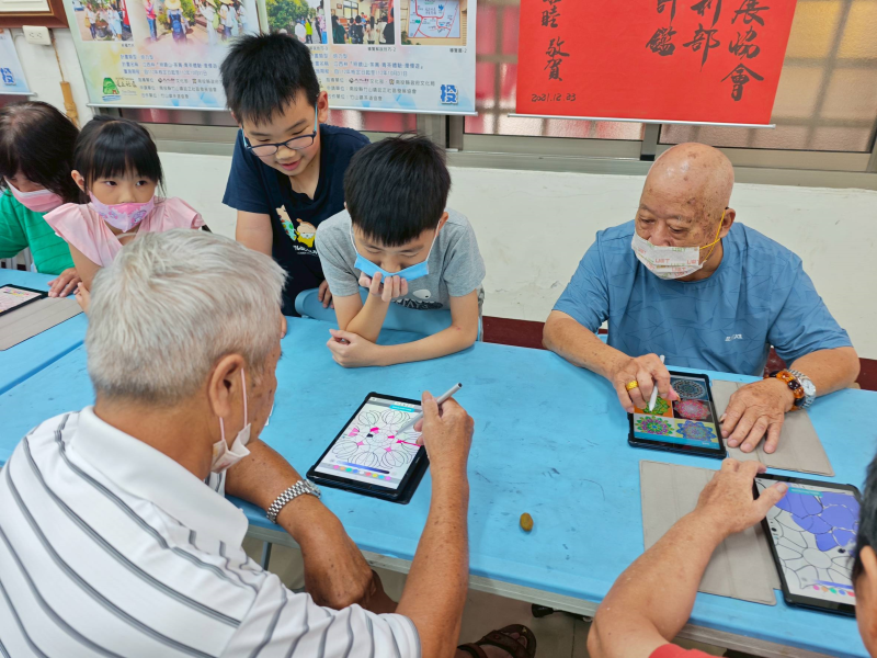 課程中，長者學員正在平板上進行數位塗鴉創作，孩子們圍在一旁協助與觀摩，孩子們熱情地提供意見與鼓勵，展現出青銀共學的互動氛圍，也讓長者在輕鬆愉快的陪伴下勇於嘗試新的科技工具。