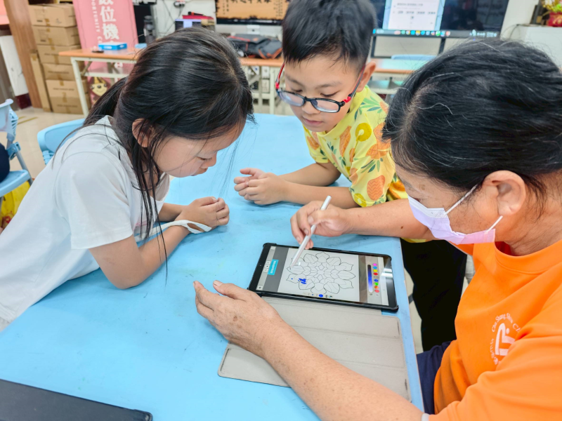 長輩正專注使用觸控筆操作平板進行繪圖練習，由於不習慣使用觸控筆來點選，兩位小朋友在旁側身觀察並適時給予協助。這樣的教學互動不僅讓孩子增進表達與教學能力，也讓長者獲得來自下一代的陪伴與鼓勵，提升自身學習自信。