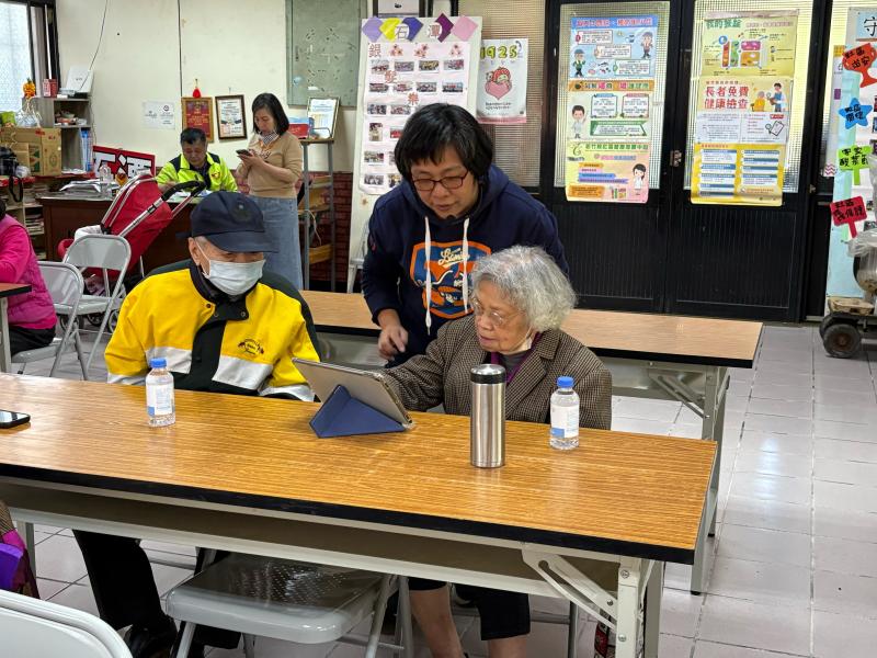 一位女性講師俯身在長桌旁，耐心地指導一位坐著的年長女性操作平板電腦，旁邊還有一位戴著口罩的年長男性一同觀看學習。