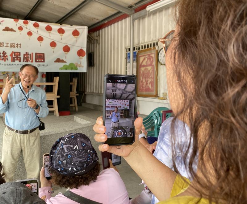 為了傳播更多工廠村的生命歷程，課程特別安排學員錄製村長的口播。照片中可見學員與村長的近距離互動，透過耆老的親口敘述，賦予了影像更強的生命力。這種「在地人說在地事」的共創模式，讓學員在學習剪輯技術的同時，也深化了對工廠村社區的情感連結與文化認