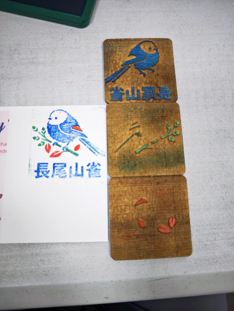 這張照片是學員超有成就感的實作成果！學員選了可愛的「長尾山雀」當主題，順利在軟體裡把圖案拆成藍色（小鳥跟文字）、綠色（樹枝）和紅色（葉子）三個印章圖層。經過雷雕製作後，大家看左邊那個蓋出來的成品，顏色清清楚楚，而且對位超級精準、完全沒歪掉！