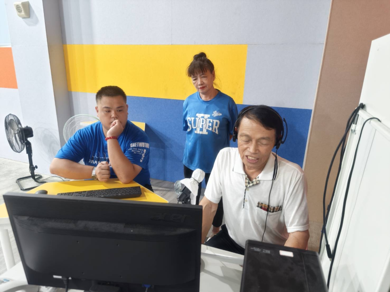 學員戴上耳機於錄音室前錄製客語音檔,旁邊有人員陪同指導